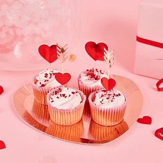 Cupcake toppers met rode hartjes en rose gouden pijltjes zitten in cupcakes en staan op een rose gouden bord in de vorm van een hart