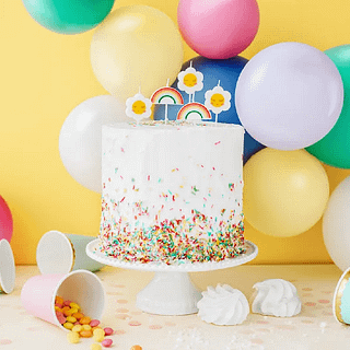 Witte taart versierd met regenboog sprinkles staat op een wite tafel voor een gele muur versierd met een ballonnenboog en kaarsjes met madeliefjes
