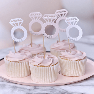 Roze cupcakes staan op een roze bordje en zijn versierd met cupcaketoppers in de vorm van diamanten ringen in het wit en roze