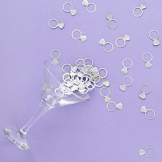 Iridescent confetti in de vorm van trouwringen liggen op een lila ondergrond naast een martiniglas