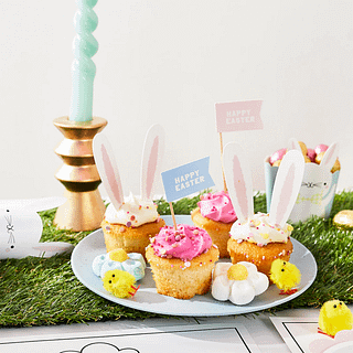 Lichtblauw bord staat op een grasmat en is gevuld met cupcakes met roze en witte creme en cupcaketoppers met vlaggetjes en konijnenoren