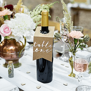 Wijnfles met bruine hanger eraan en de witte tekst table one staat op een witte tafel voor een bloemstuk