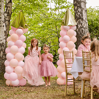 Twee ballonnenbogen die lijken op kasteeltorens staan in een bos en er lopen twee meisjes doorheen in roze jurken