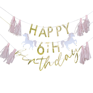 Personaliseerbare slinger met gouden letters, witte paarden en roze franjes hangt in een bos voor een zachtroze ballonnenboog
