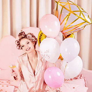 Vrouw met rollers in haar haren zit op bed naast een tros roze ballonnen