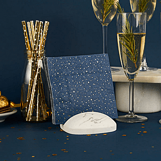 Donkerblauwe servettenmet gouden sterren, stippen en streepjes staan in een marmeren houder naast een champagneglas en gouden rietjes