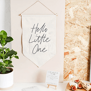 Stoffen doek met de tekst hello little one hangt naast een plant en voor een witte muur