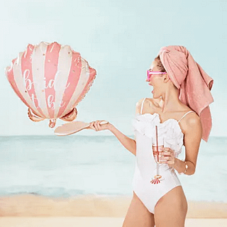 vrouw zit op strand met folieballon in de vorm van een schelp in het roze en beige