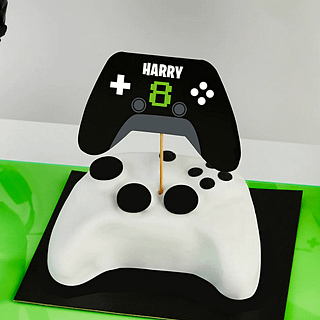 personaliseerbare taart topper in de vorm van een zwarte controller met stickervellen in het groen en wit in een witte taart in de vorm van een playstation controller