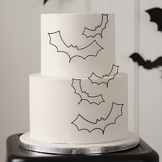 Zwarte, metalen vleermuizen zitten aan de zijkant van een witte taart met twee verdiepingen die staat op een zilver plateau en zwart krukje