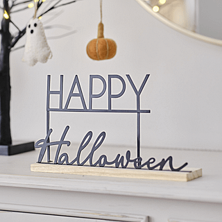 Metalen bordje met zwarte tekst happy halloween op een houten plank staat op een witte kast naast een lichtgevende boom met pompoenen eraan hangen
