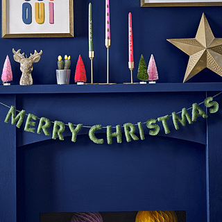 Groene slinger van stof met de tekst merry christmas hangt voor een marinablauwe schouw gevuld met kleurrijke kerstversiering