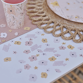 Perzik en roze confetti in de vorm van bloemen en de tekst happy birthday ligt op tafel naast een rieten placemat