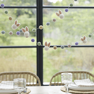 Slinger met pom poms en konijnenoren hangt boven een houten tafel voor een raam met zwarte kozijnen