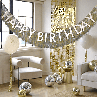 Gouden tassel slinger met de nude tekst happy birthday hangt in een woonkamer versierd met taupe ballonnen en een gouden backdrop