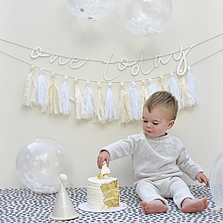 Jongetje zit op zijn eerste verjaardag bij een taart tussen de witte en creme versiering met confetti ballonnen en een tassel slinger