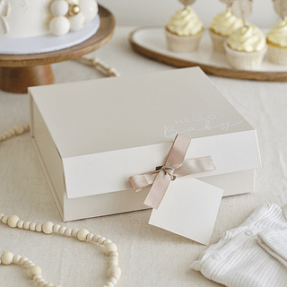 Creme cadeau doos met licht bruin lint en de tekst hello baby ligt op tafel naast een houten kralen slinger