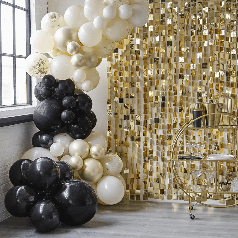 Zwarte, champagnegouden en nudekleurige ballonnenboog staat voor een gouden backdrop met metalen vierkantjes bij een raam met zwarte kozijnen