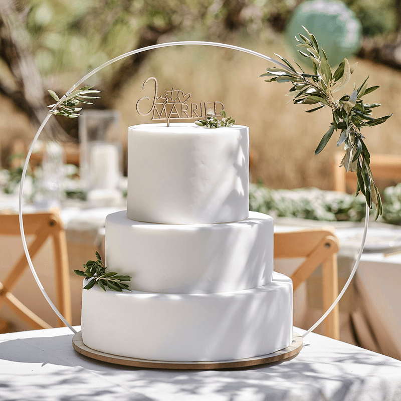 Witte taart van drie verdiepingen staat op een witte tafel op een houten plateau met een witte boog eromheen en is versierd met groene takjes en een houten taart topper