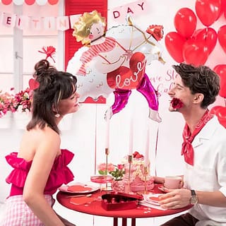Man en vrouw zitten aan een rode tafel met roze servies en achter hen zweeft een folieballon in de vorm van Cupido