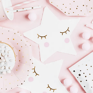 Witte servet in de vorm van een ster met gouden wimpers ligt op een lichtroze achtergrond bedekt met pompoms en marshmallows