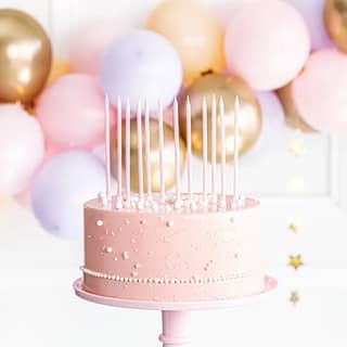 Zes lange lichtroze taartkaarsen op een roze taart met ballonnen erachter