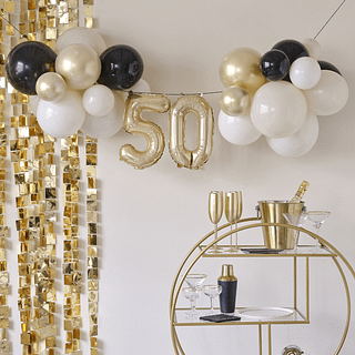 Ballonnenslinger voor 50 jaar in het goud, nude en zwart hangt boven een gouden karretje met champagneglazen en een cocktailshaker en voor een gouden backdrop met vierkantjes