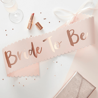 Lichtroze sjerp met de rosé gouden tekst 'Bride to be' ligt naast een glas champagne en een roze lippenstift
