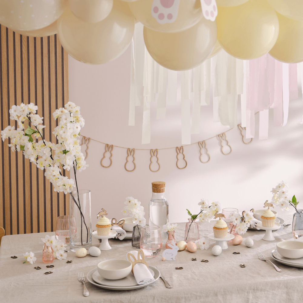 Houten tafel met ivoorkleurig tafelkleed en paasversiering in verschillende pasteltinten bestaande uit ballonnen, eieren en een houten slinger
