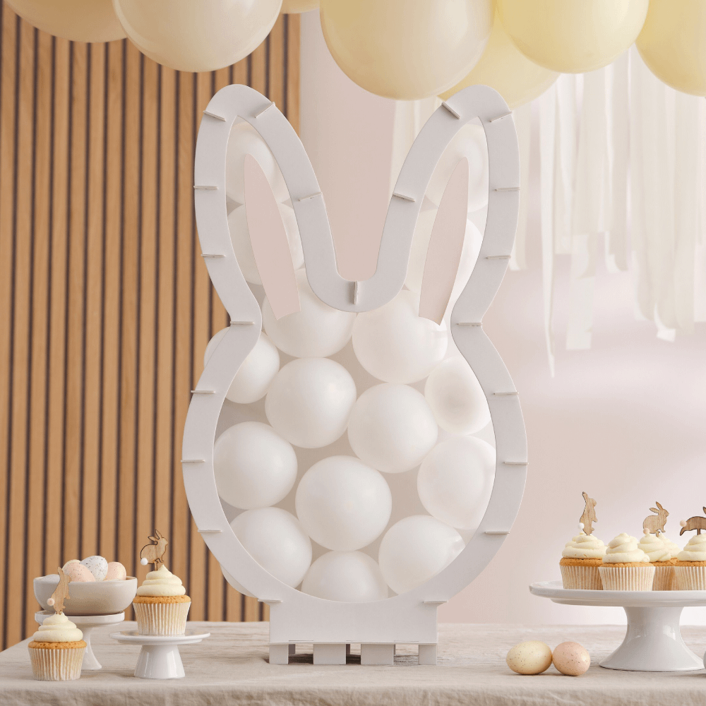 Ballonnenstandaard van een wit konijn staat op een tafel naast cupcakes en voor een schaal met gekleurde paaseieren