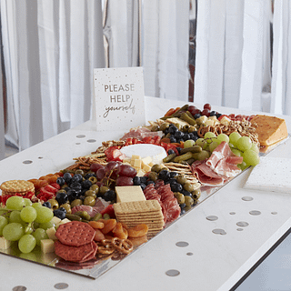 Zilveren hapjesplank op een witte tafel bedekt met zilveren confetti en pretzels, druiven, kaas en andere hapjes