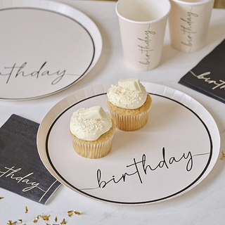 Nudekleurig bordje met de zwarte tekst 'birthday' en een zwarte rand staat naast gouden confetti, een zwarte servet en is gevuld met cupcakes